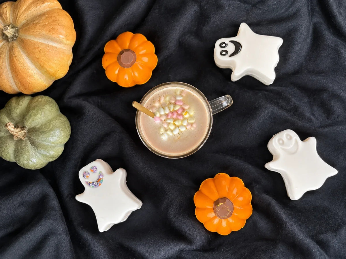 Kopp med kakao og marshmallows omgitt av hvite spøkelseskakaobomber og oransje gresskarkakaobomber på mørkt stoff.