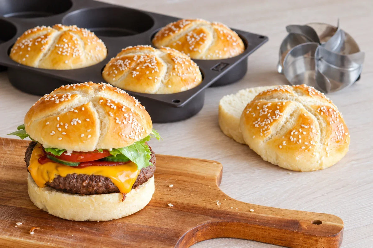 Hamburger med hjemmelaget sesambrød på trefjøl, med flere hamburgerbrød ved siden av.