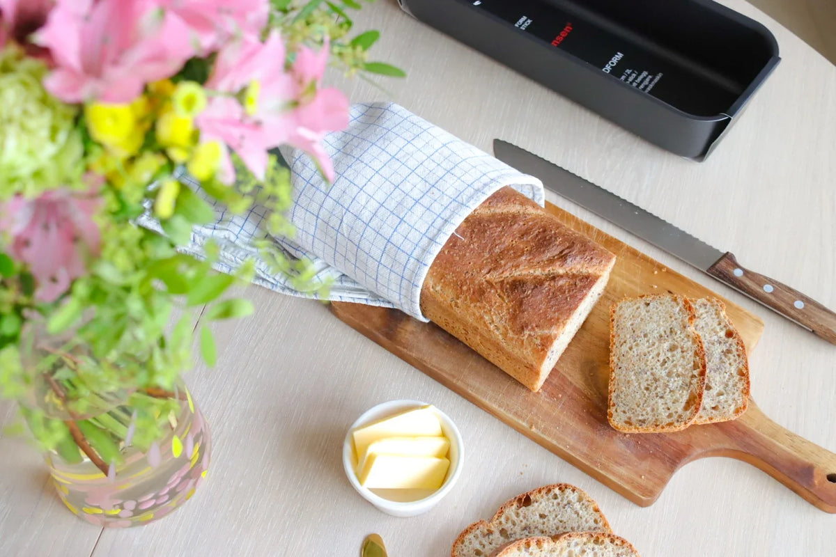 Halvgrovt brød med to skiver på en trefjøl, med smør, blomster og en brødform i bakgrunnen.