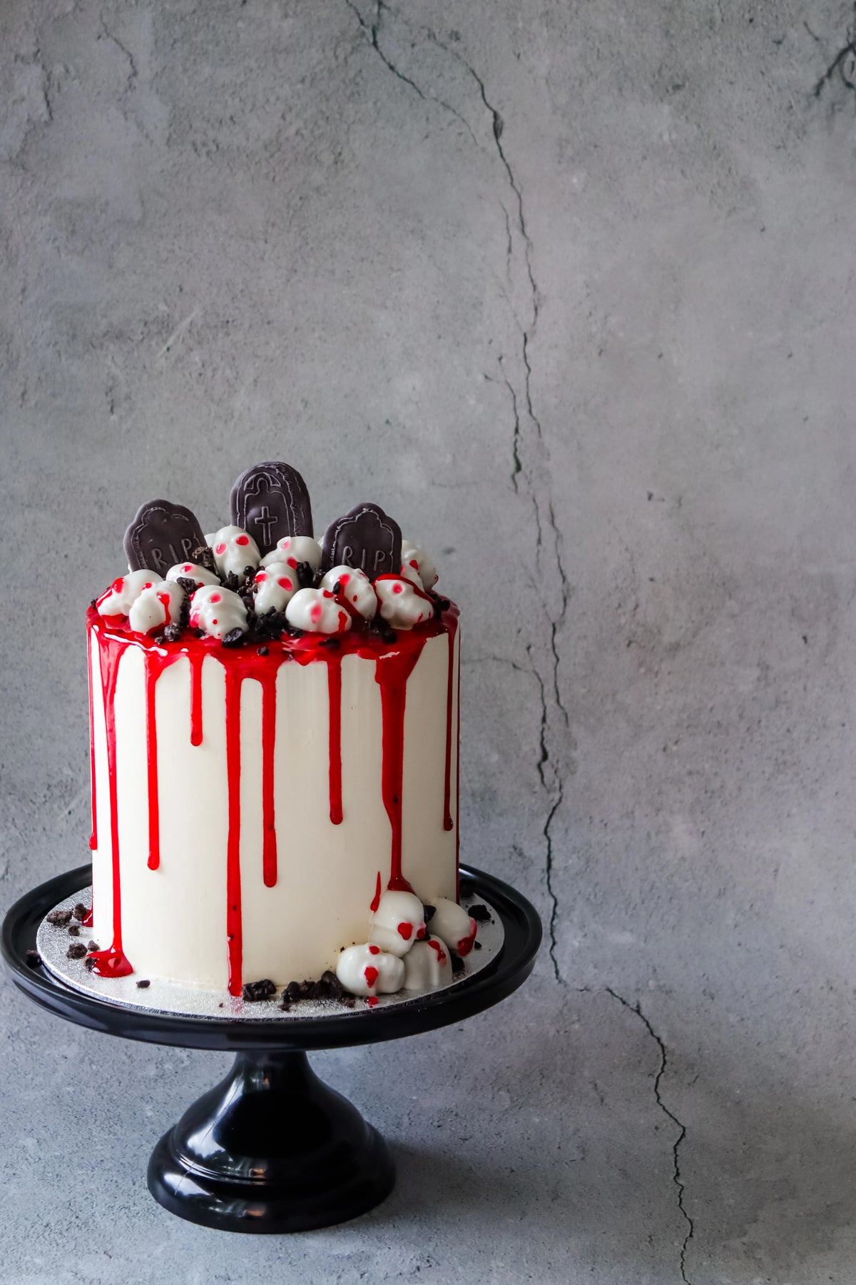 Halloweenkake med hodeskaller, gravsteiner og rød glasur på svart stettfat foran grå betongvegg.